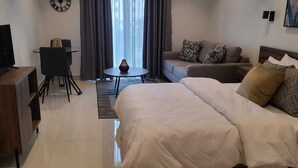 A hotel room with a bed, a sofa, a TV, a small table, and a chair. - Belleson Homes The Lennox (Accra)