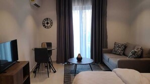 A hotel room with a TV on a wooden stand, a sofa, a small round table with a vase, a chair, and a clock on the wall. - Belleson Homes The Lennox (Accra)