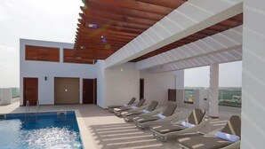 A rooftop pool area with a wooden pergola, lounge chairs, and a clear view of the city skyline. - Belleson Homes The Lennox (Accra)