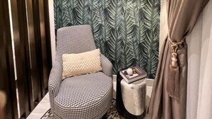 A room with a checkered armchair, a small ottoman, a patterned rug, a decorative wall with a leaf pattern, and a gold-framed window. - Belleson Homes The Lennox (Accra)