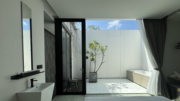 A modern bedroom with a large bed, a built-in sink, a mirror, and a sliding glass door leading to a balcony with a potted plant.