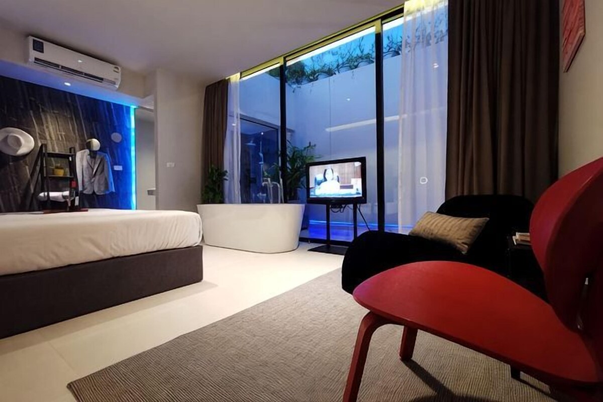 Quarto de hotel moderno com cama, uma cadeira vermelha, televisão e uma janela grande.