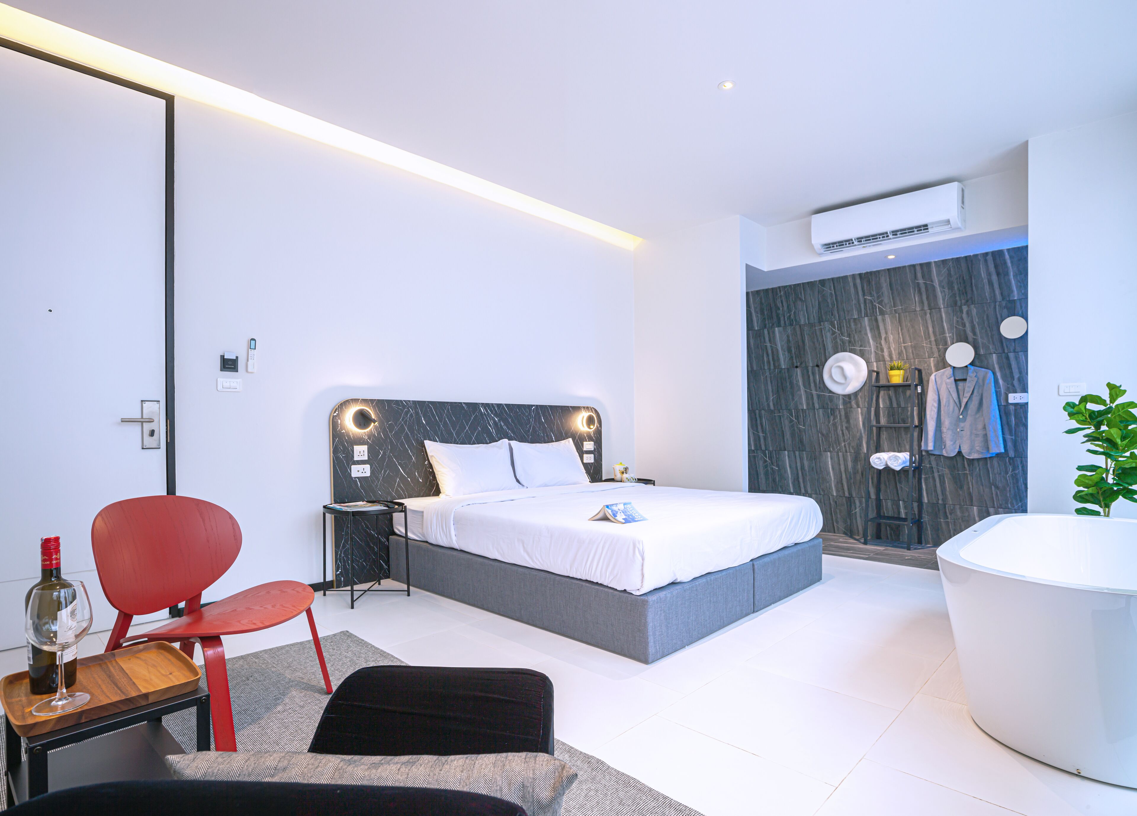 A modern hotel room with a bed, a red chair, a small table with a wine bottle, and a large white bathtub.