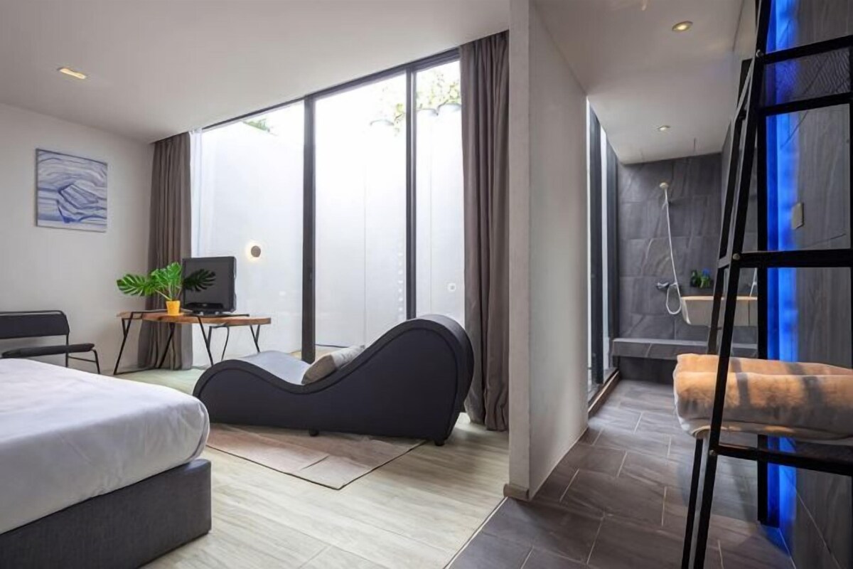Quarto de hotel moderno com cama, escrivaninha, poltrona, chuveiro e varanda com janelas amplas.
