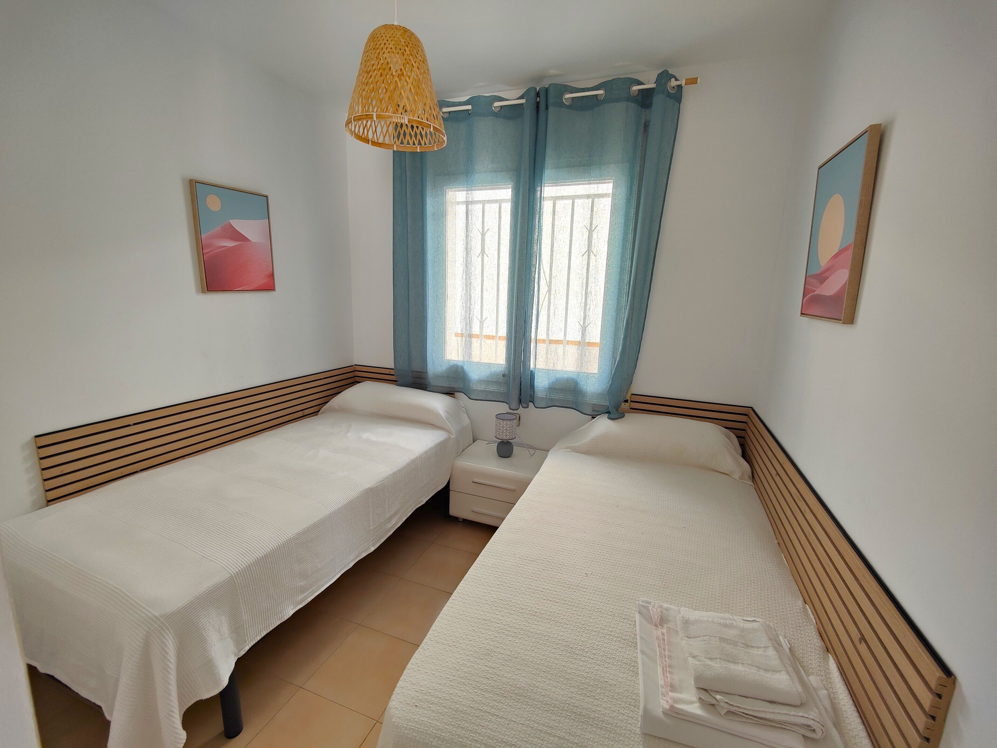 Une chambre avec deux lits simples, une fenêtre avec des rideaux bleus, un luminaire suspendu en rotin et deux tableaux encadrés accrochés aux murs.