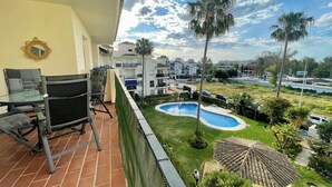 A balcony with a glass table and chairs, overlooking a swimming pool and a grassy area with palm trees. - Stunning 2Bed Apartment in Marbella - Pto Banus (Marbella)