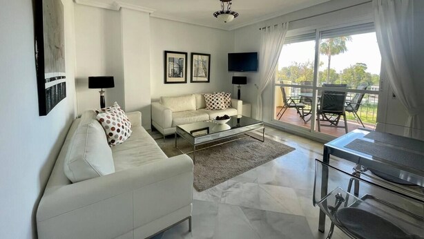 A modern living room with a white sofa, a glass coffee table, and a TV. There is a balcony with outdoor furniture. - Stunning 2Bed Apartment in Marbella - Pto Banus (Marbella)
