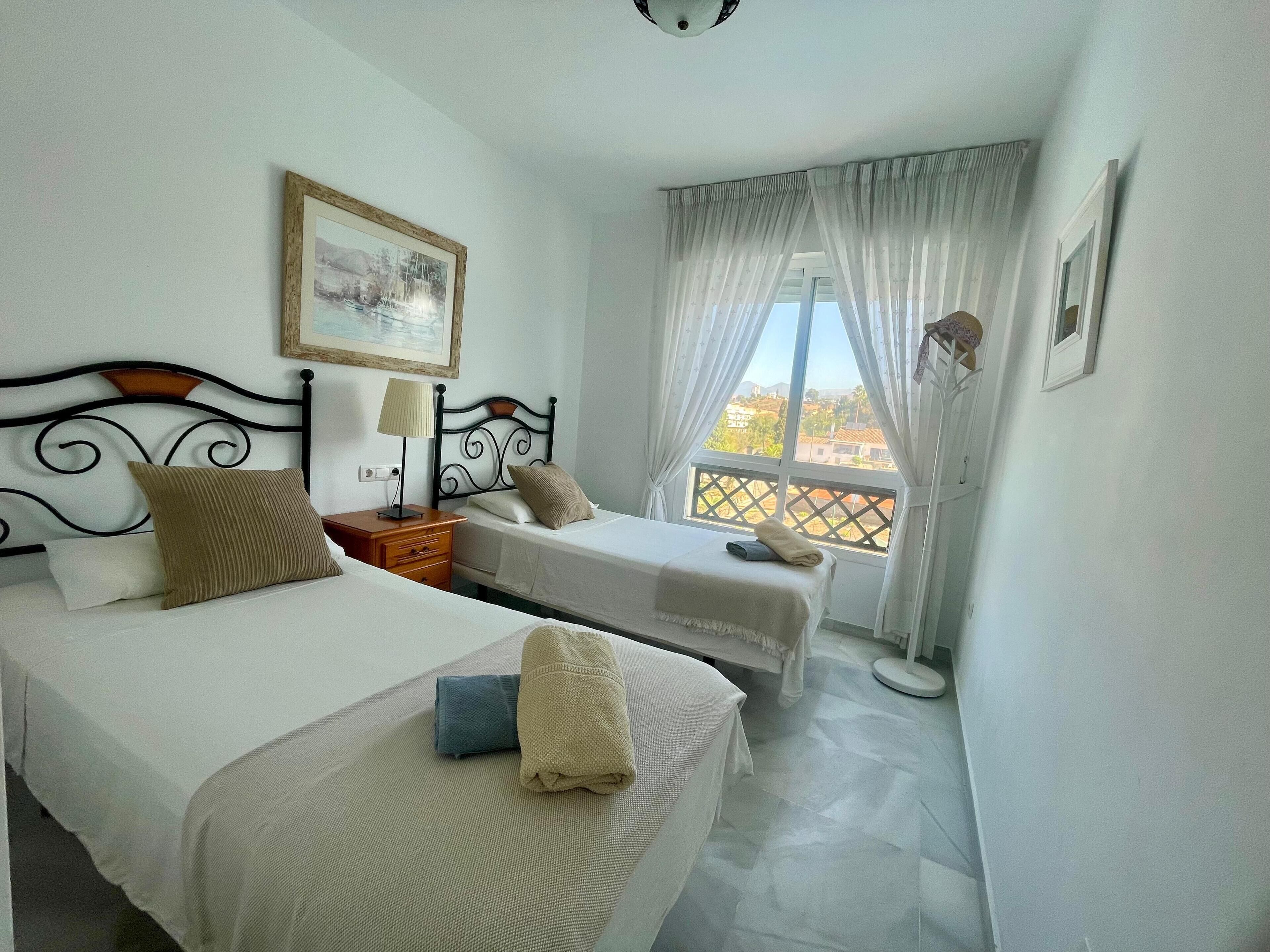 A hotel room with two beds, a nightstand, a lamp, and a window with a view of the city.