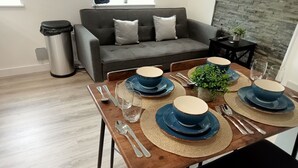 Ein Essbereich mit einem Holztisch, gedeckt für vier Personen, inklusive blauer Teller, Besteck und klaren Gläsern. Im Hintergrund steht ein graues Sofa und ein kleiner Beistelltisch mit einer Pflanze.