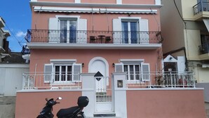 Un immeuble de deux étages avec une façade rose, des volets blancs et un balcon doté de rampes en métal.