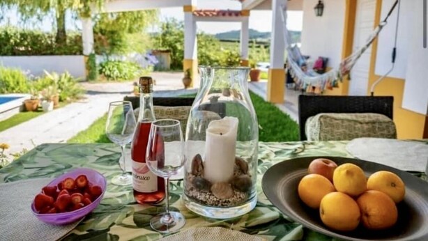 Une table avec un chandelier en verre, un bol de fraises, une bouteille de vin et une assiette d’oranges.