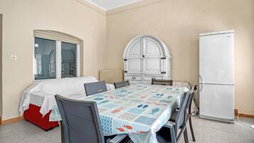 Una habitación con cama, una mesa con sillas, un refrigerador y un ventanal con cortinas.