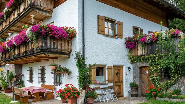 A traditional chalet with a stone facade, wooden balconies with flower boxes, and a stone patio with outdoor seating.