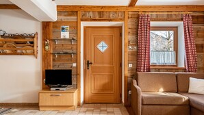 A wooden interior with a wooden door, a television on a wooden stand, a brown sofa, and a window with curtains.