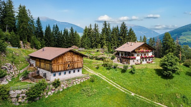 Un village de montagne aux maisons traditionnelles en bois, avec un puits en pierre et une vue dégagée sur la forêt et les montagnes environnantes.