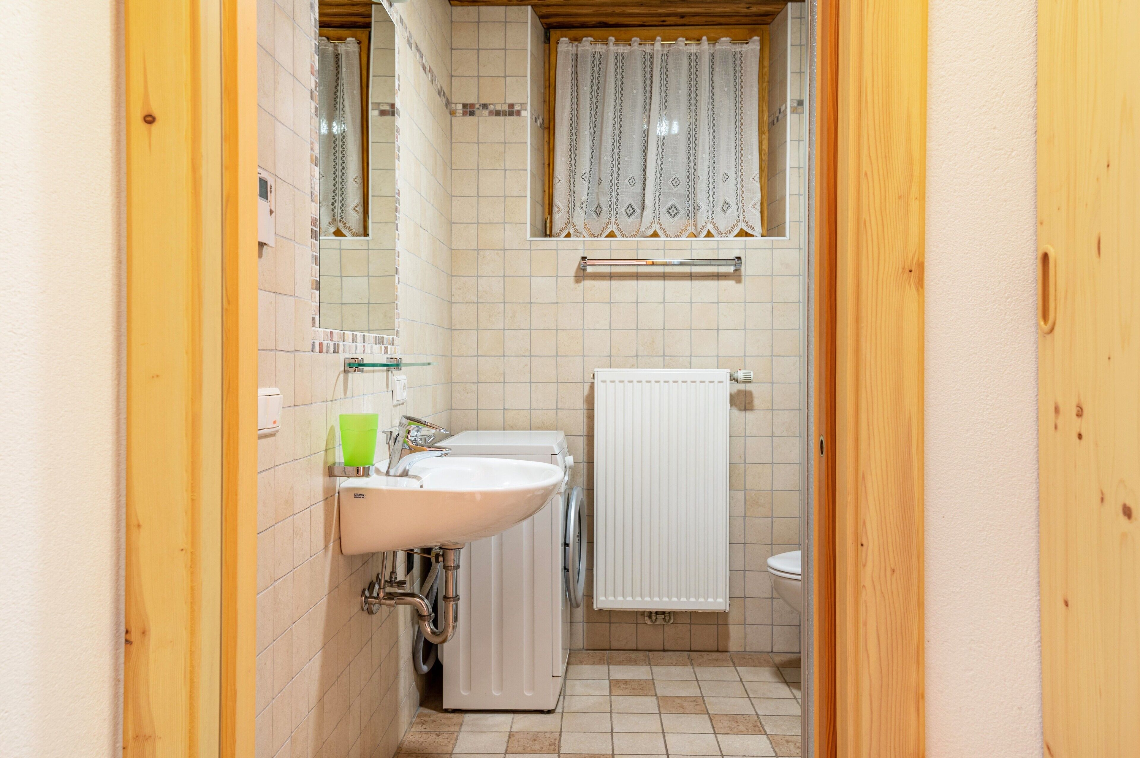 Un bagno con lavandino, specchio, finestra ornata da tende di merletto, porta asciugamani e servizi igienici.