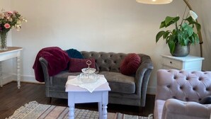 Un salon avec un canapé gris, une table d’appoint blanche, une petite table blanche recouverte d’une nappe en dentelle, un plaid rouge et une plante verte.