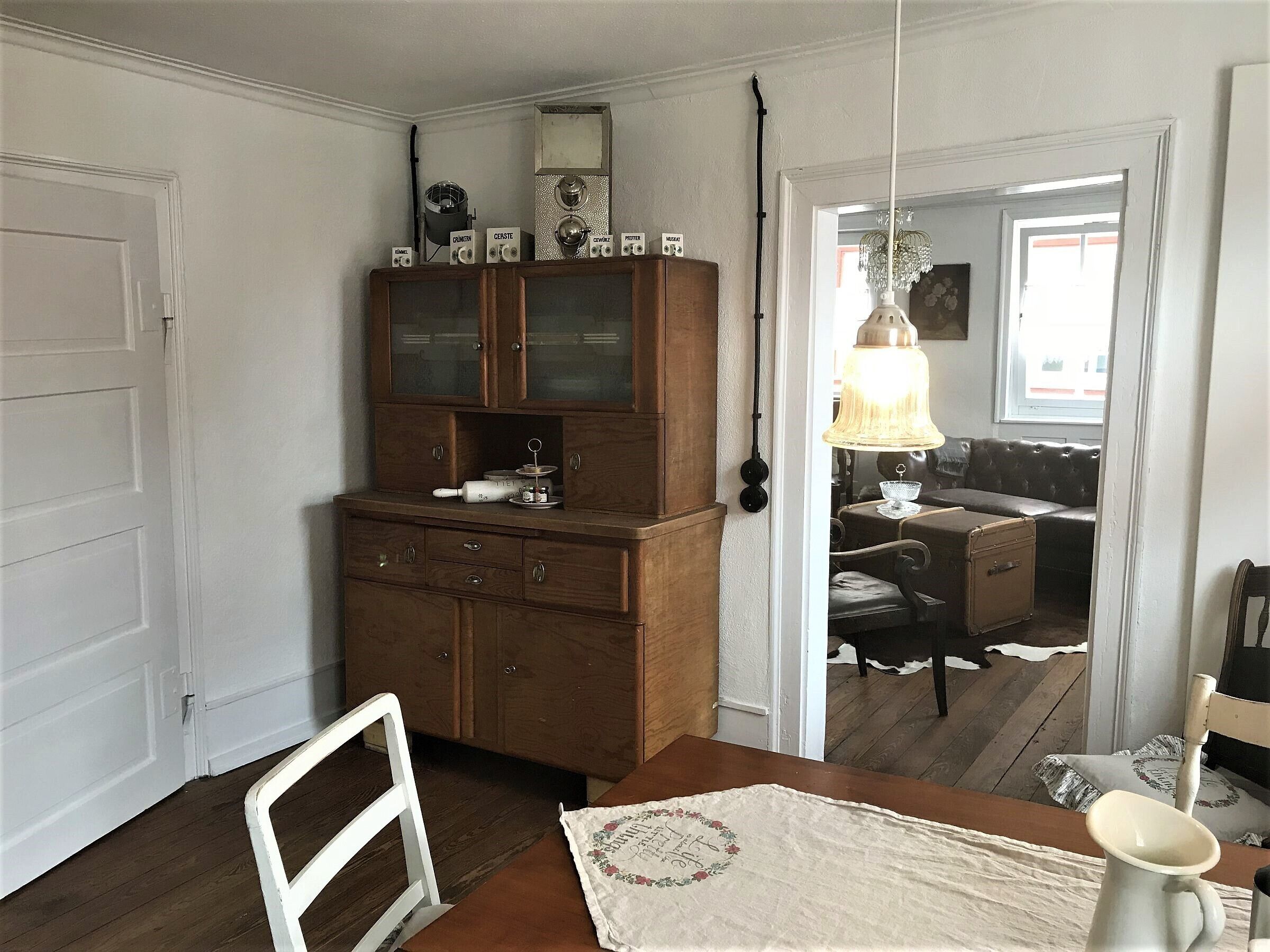 Une pièce comprenant un meuble en bois, une table à manger avec un set de table, une chaise blanche, une valise ancienne et un lustre.
