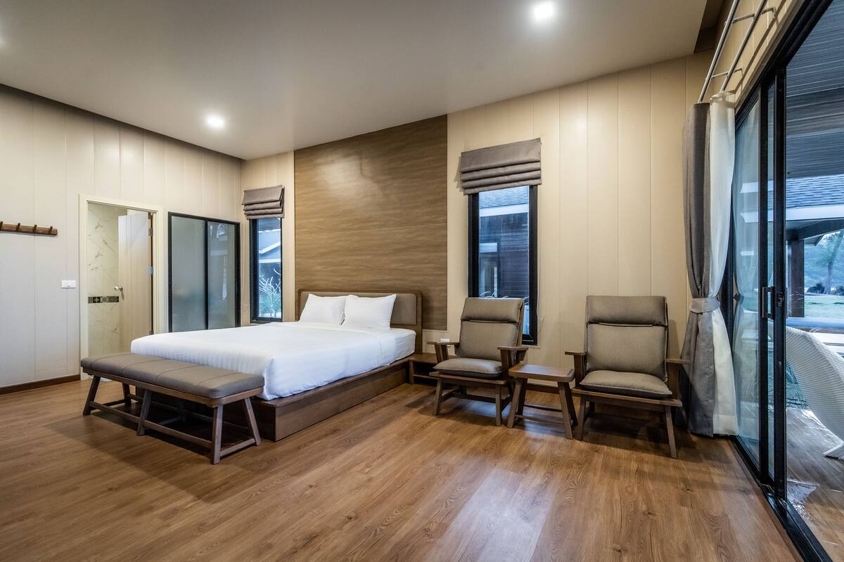 A modern bedroom with a large bed, two chairs, a bench, and a sliding glass door leading to a balcony.