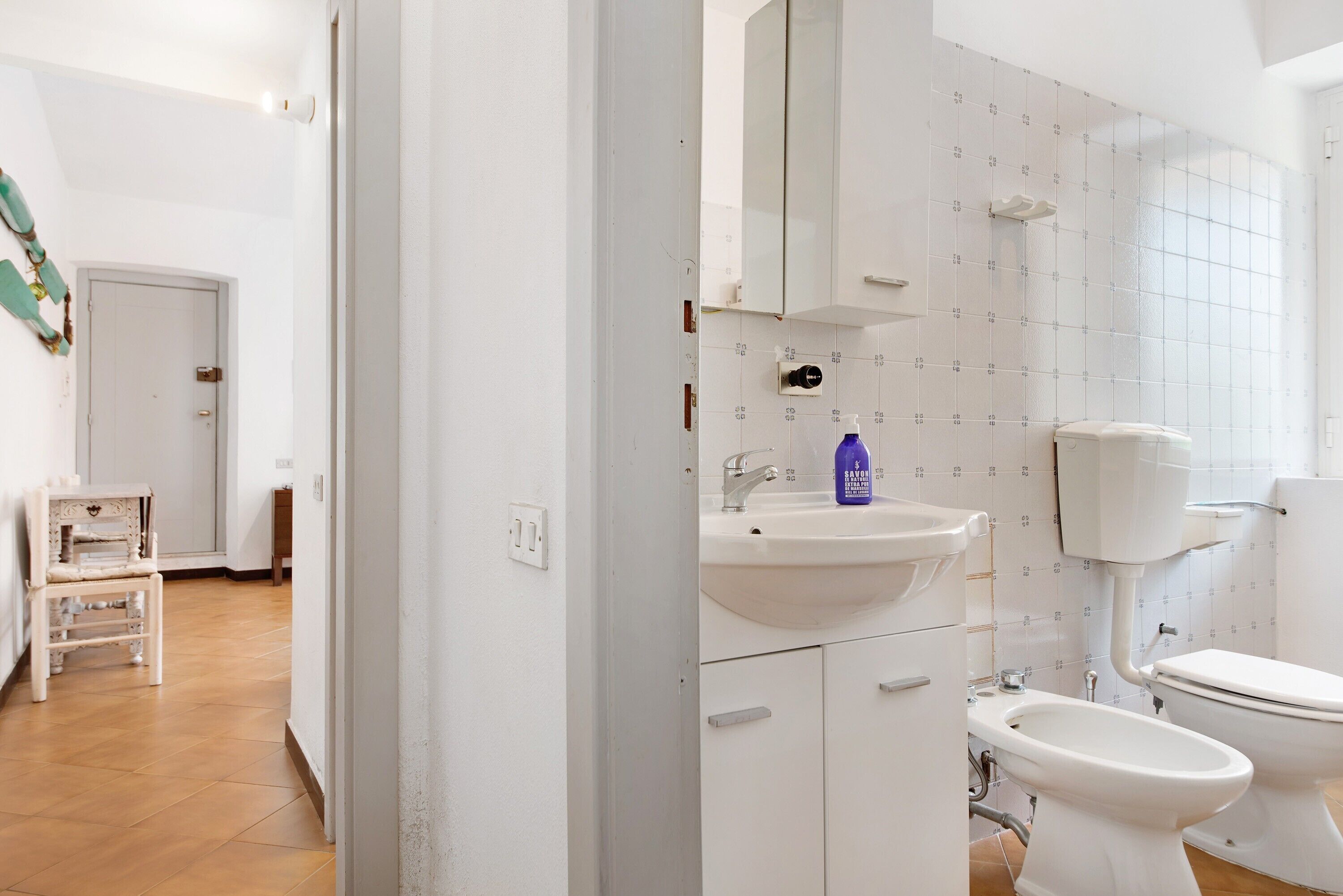 Een badkamer met een wit wastafelmeubel, toilet en bidet, een spiegel boven het wastafelmeubel, en een betegelde wand.