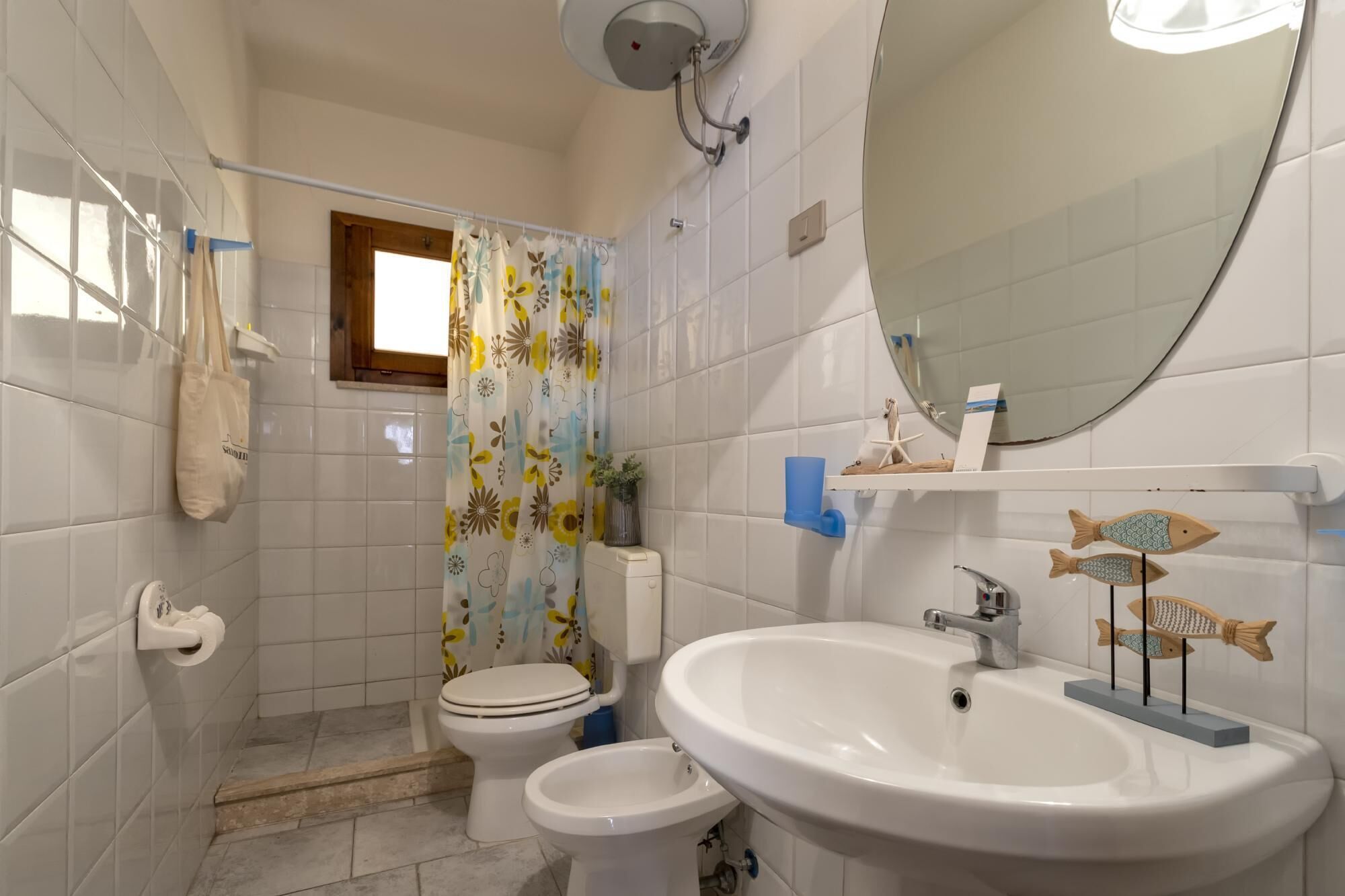 Kylpyhuoneessa on valkoiset laatat, wc-istuin, bidee, pesuallas ja kukkakuviollinen suihkuverho.