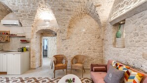 Un interior rústico de piedra con cocina, dos sillones de mimbre, un sofá y un tapete estampado.