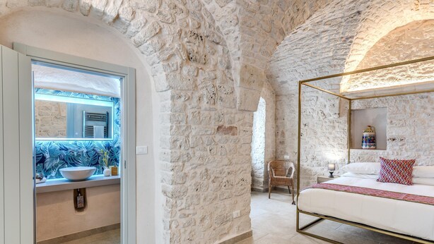Un dormitorio con pared de piedra, una cama con estructura dorada, una ventana que da a un baño, y una silla cerca de la ventana.
