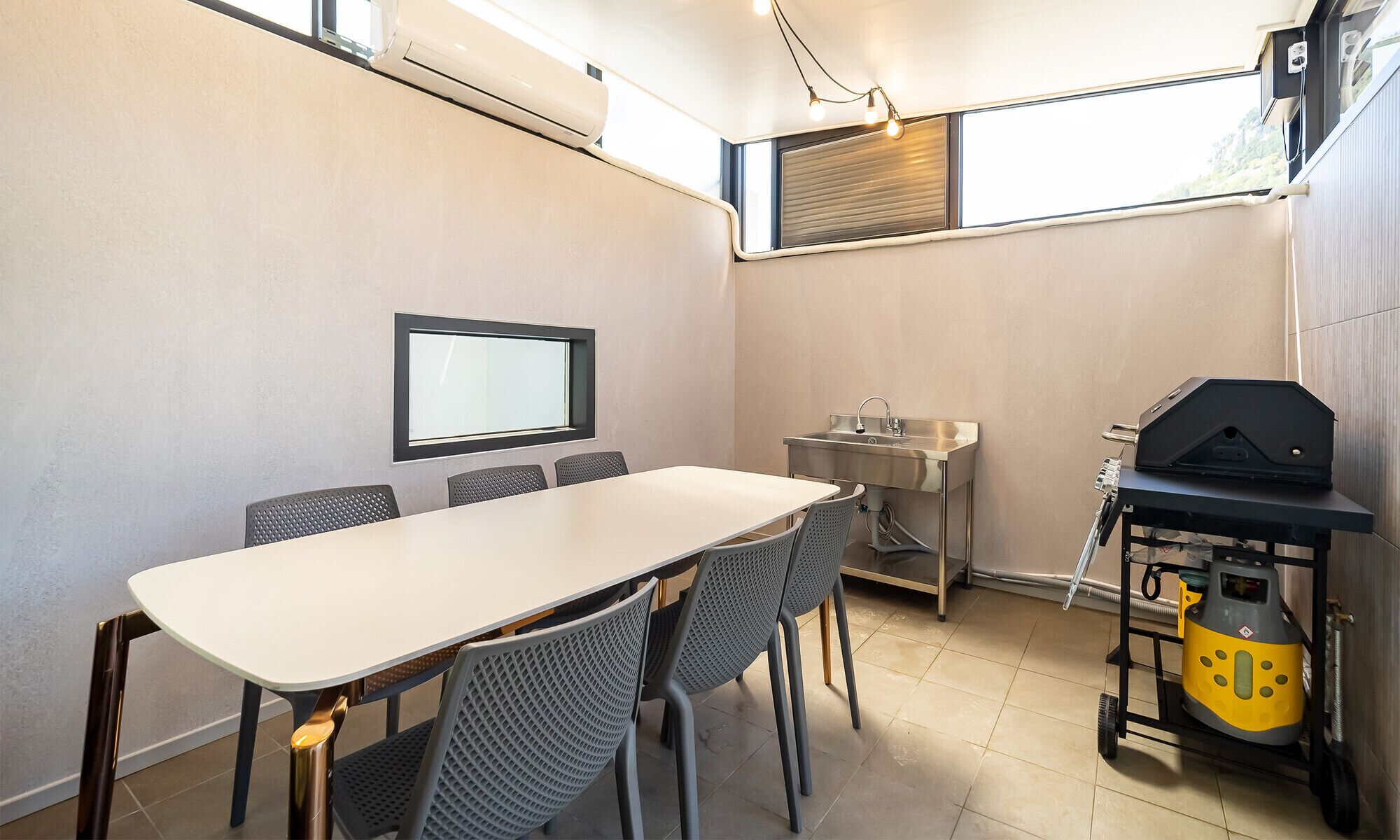Una cocina compacta con una mesa de comedor, sillas, un lavamanos integrado, una ventana con persianas y una parrilla portátil.