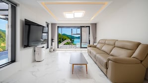 A modern living room with a large sofa, a flat-screen TV, a coffee machine, and a view of a scenic landscape through large windows. - Baekya Pool Villa & Kids (Yeosu)