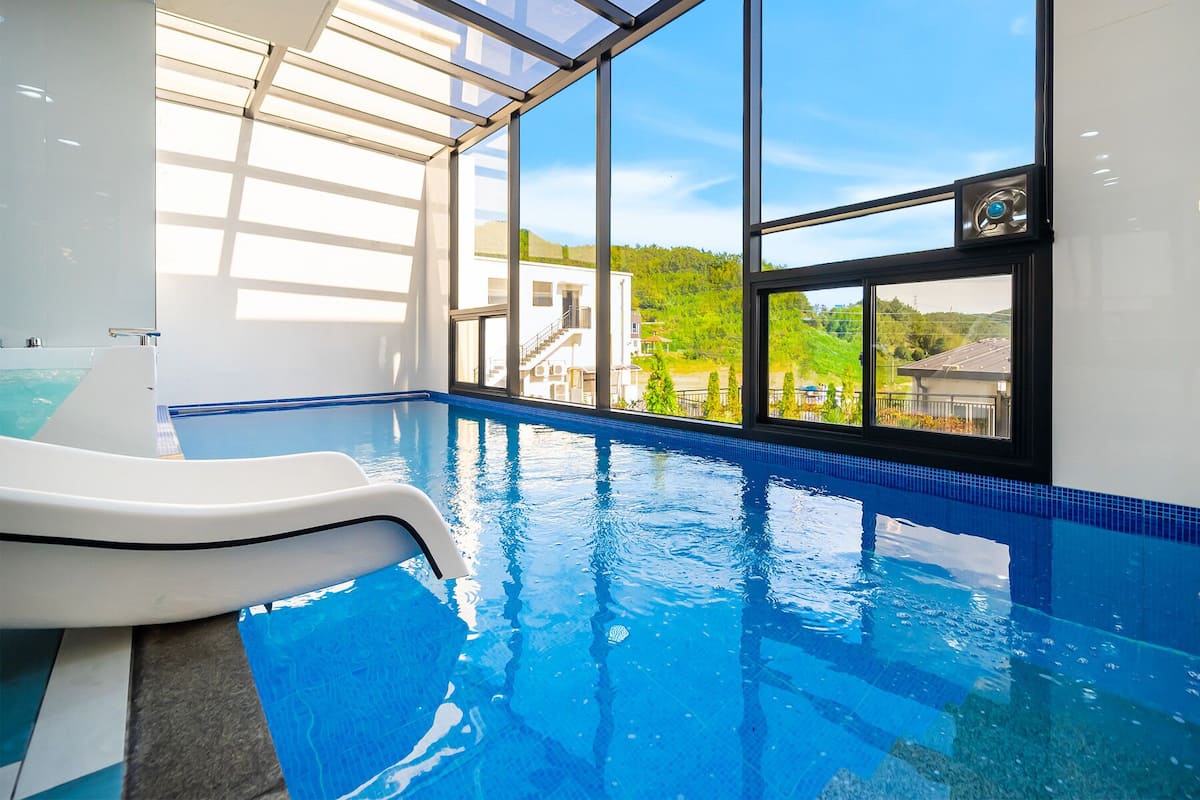 An indoor swimming pool with clear glass walls, a view of a residential area, and a water slide.