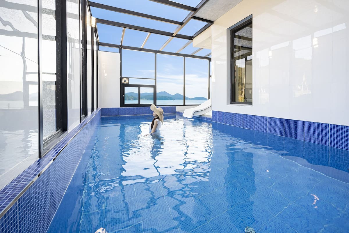 A large indoor swimming pool with clear blue tiles, a glass ceiling, and a view of mountains.