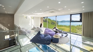 A modern hotel room with a glass floor, a large window offering a scenic view of a lake and mountains, and a staircase leading to an upper level. - Baekya Pool Villa & Kids (Yeosu)