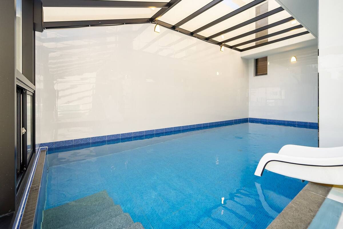 A clear indoor swimming pool with a blue tiled edge, a white poolside area, and a glass ceiling.