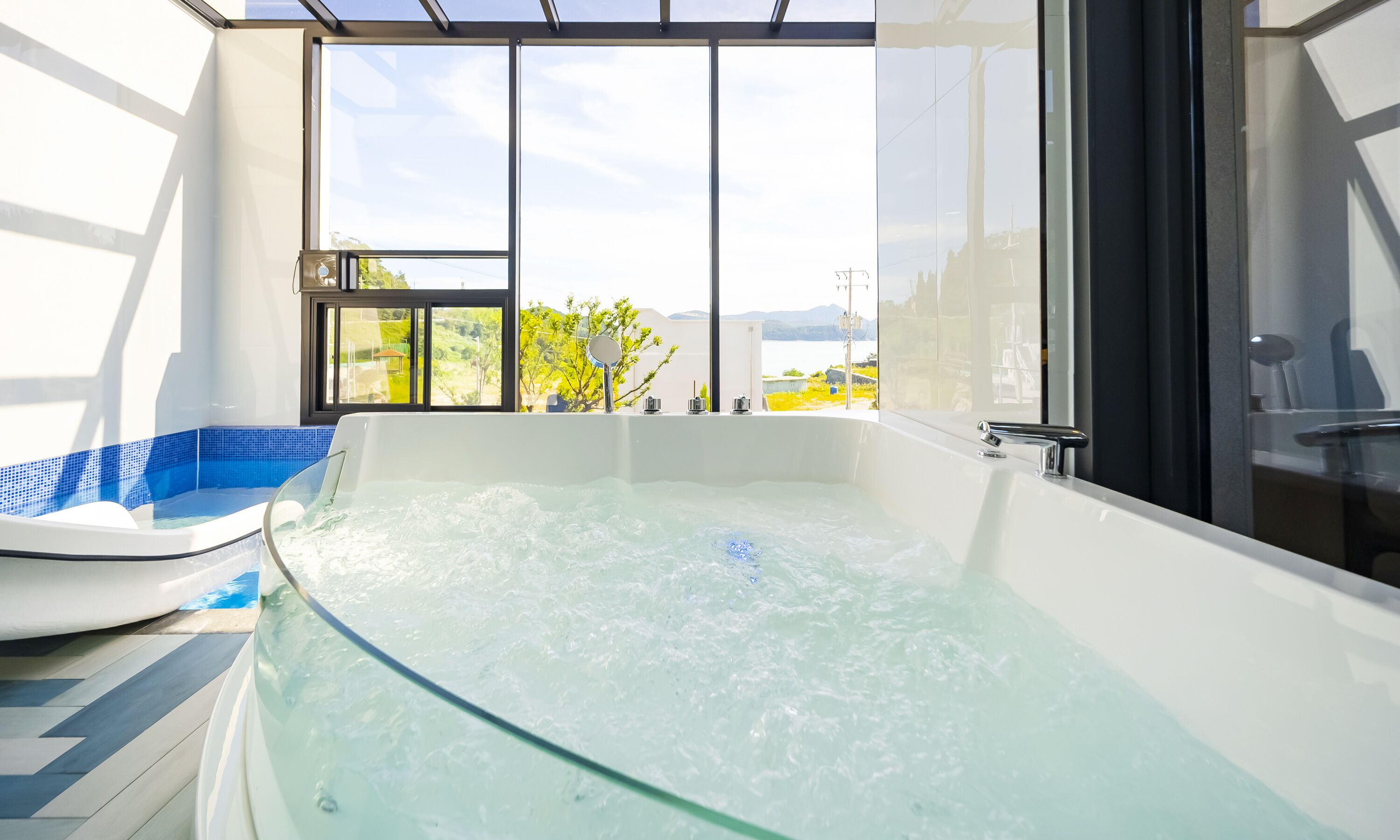A modern bathroom with a glass-enclosed jacuzzi, a large window, and a view of the outdoors.