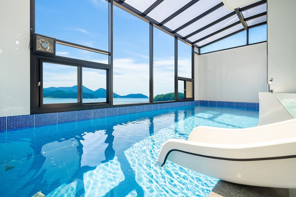A modern indoor swimming pool with clear blue water, large windows offering a view of distant mountains, and a white lounge chair.
