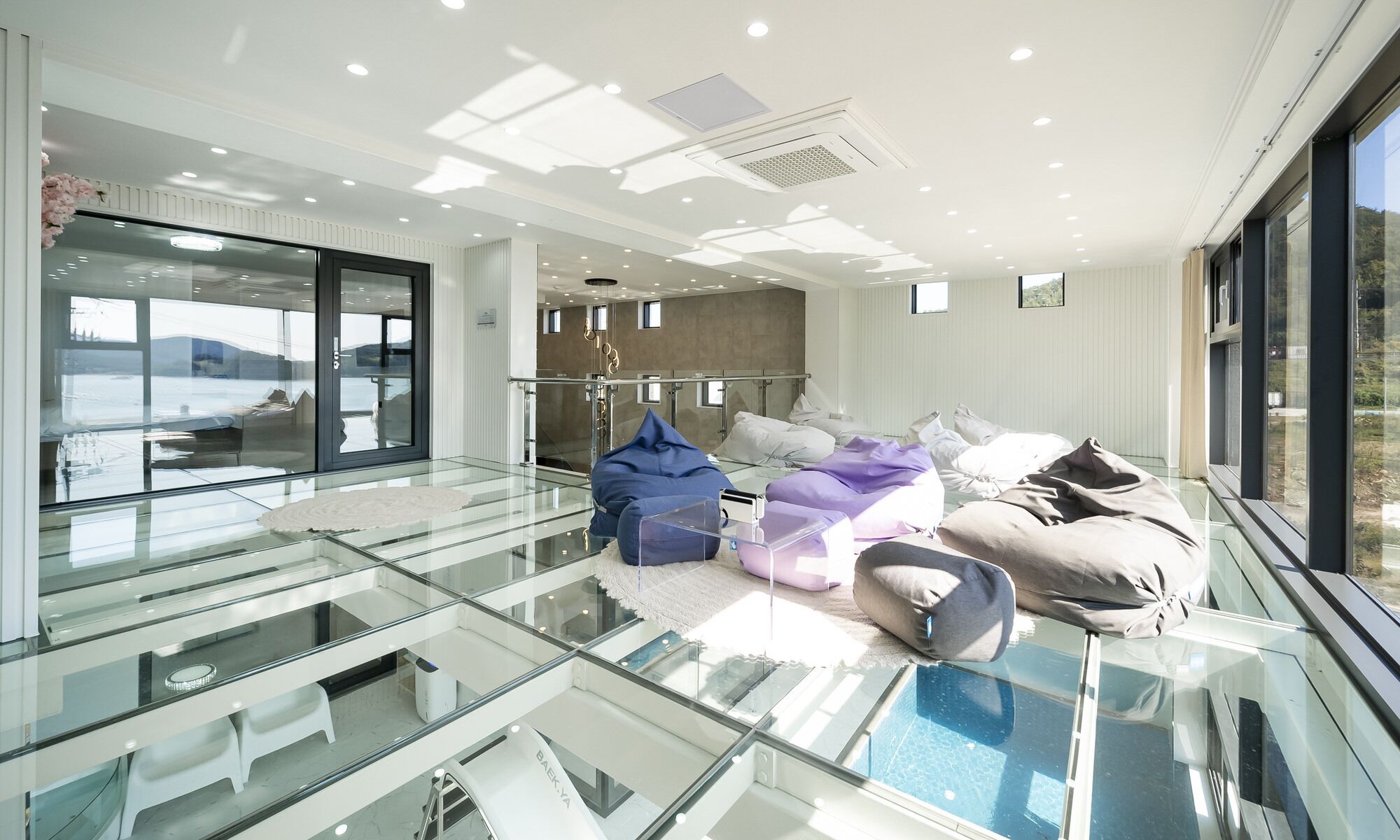 A modern hotel lobby with a glass floor leading to a pool area, a seating area with cushions, and a view of the sea.