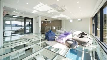 A modern hotel lobby with a glass floor leading to a pool area, a seating area with cushions, and a view of the sea.
