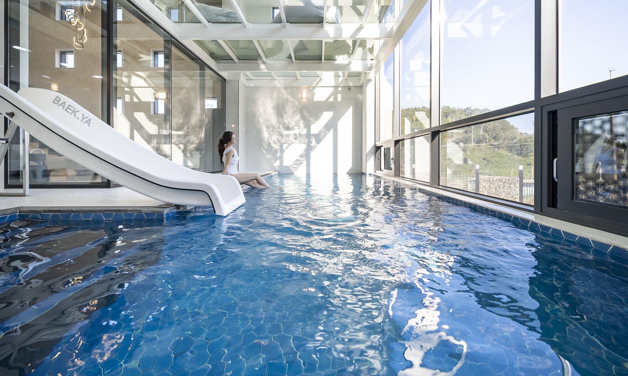 An indoor swimming pool with a slide, large windows, and a view of the outdoors.