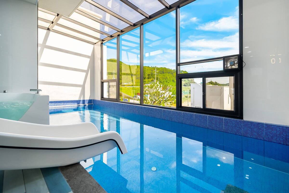 A modern indoor swimming pool with clear glass walls, a white jacuzzi, and a view of greenery outside.