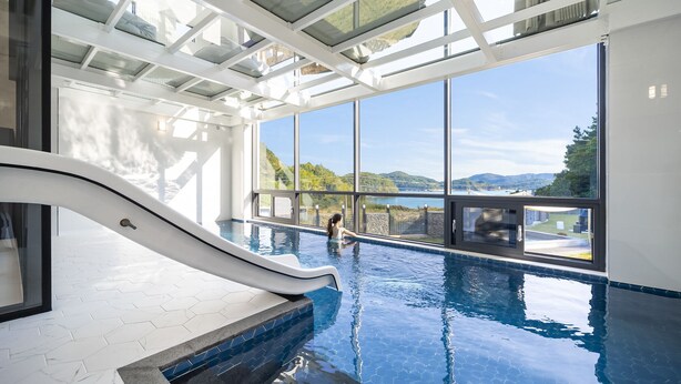 An indoor swimming pool with a glass ceiling, a slide, and a view of the landscape. - Baekya Pool Villa & Kids (Yeosu)