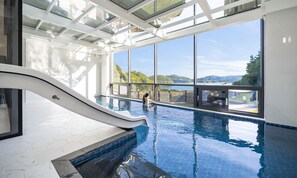 An indoor swimming pool with a glass ceiling, a slide, and a view of the landscape. - Baekya Pool Villa & Kids (Yeosu)