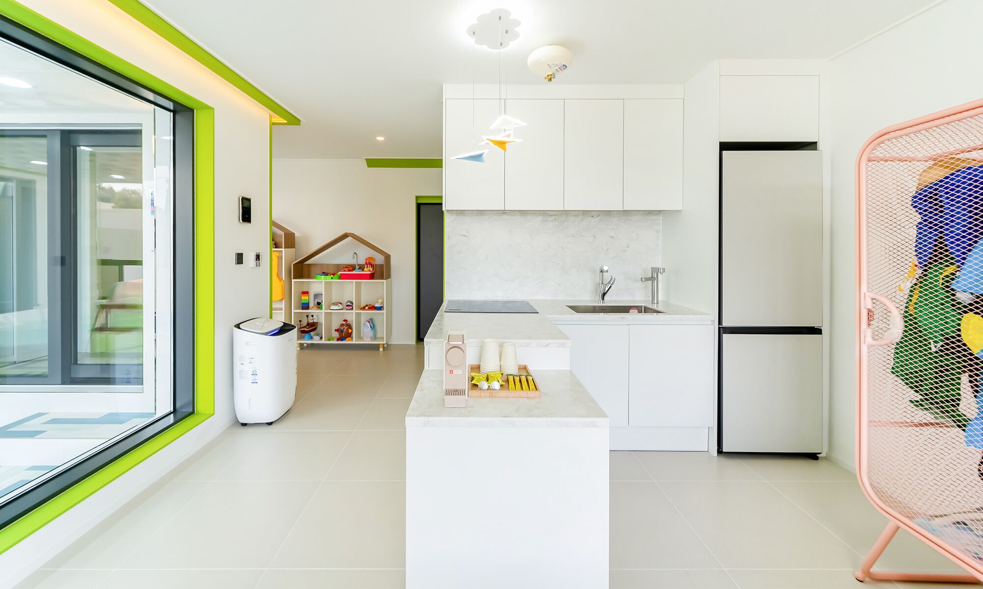 A modern kitchen with white cabinets, a built-in oven, and a sink. There is a play area with toys and a small house model.