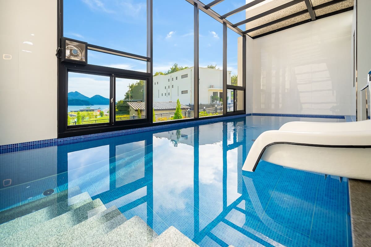 A clear indoor swimming pool with a view of mountains and buildings through large windows.