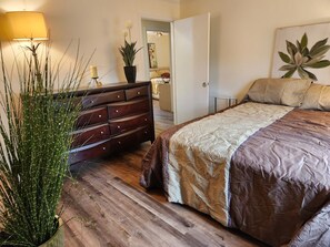 A bedroom with a wooden floor, a bed with a patterned bedspread, a dresser, a potted plant, and a lamp. - Byrd Nest @ Coventry · 3BR/2BA Home |Near Hospital, Downtown & US64/I-95 (Rocky Mount)