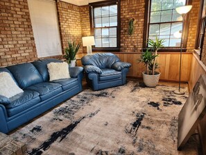 A room with a blue leather sofa, a blue armchair, a patterned rug, and a potted plant. - Byrd Nest @ Coventry · 3BR/2BA Home |Near Hospital, Downtown & US64/I-95 (Rocky Mount)