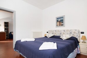 Un letto rifatto con copriletto blu e asciugamani bianchi, una testiera floreale, un quadro appeso al muro e una lampada da comodino.