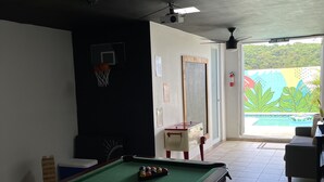 Une table de billard avec des billes, une table de baby-foot, un panier de basket et une fenêtre donnant sur un paysage verdoyant.