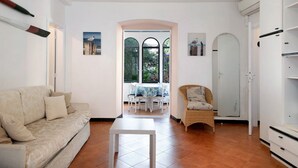 Een woonkamer met een bank, een tafel, een rieten stoel, een garderobe en uitzicht door een raam.