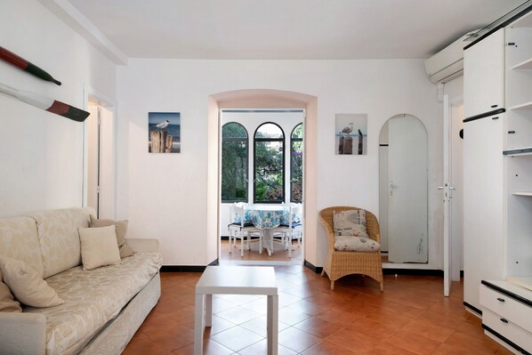 Un salón con sofá, una mesa, una silla de mimbre, un armario y vistas a través de una ventana.