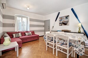 Een woonkamer met een rode bank, een eettafel met stoelen, een raam met gordijnen en een muur met een afbeelding van nautische thema's.